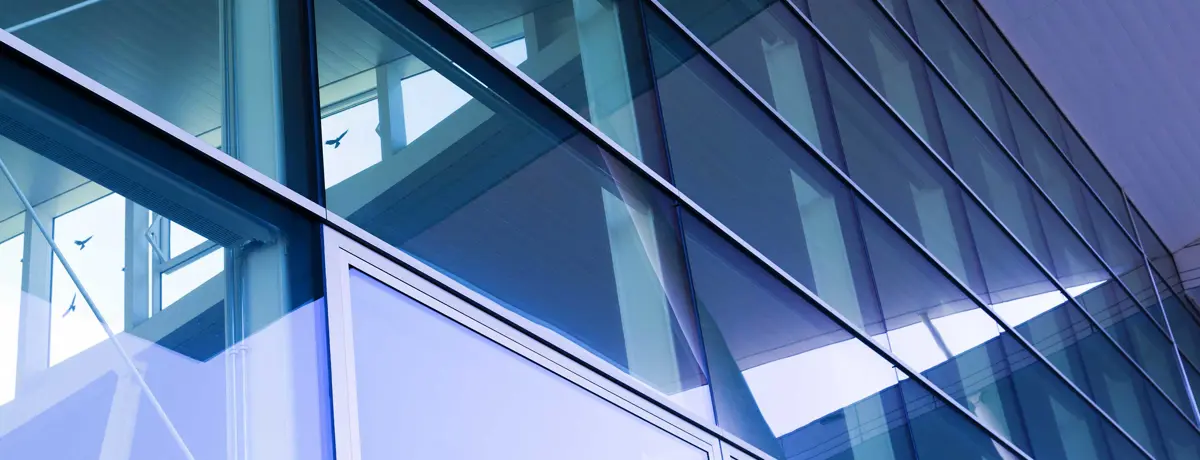 Detail van een moderne glazen gevel met grote raampartijen en een regelmatig raster van metalen profielen. In het glas zijn delen van het gebouw en de omgeving weerspiegeld. FR: Détail d’une façade-rideau moderne composée de grandes baies vitrées et d’une trame régulière de profilés métalliques. Le vitrage reflète certaines parties du bâtiment et de son environnement.